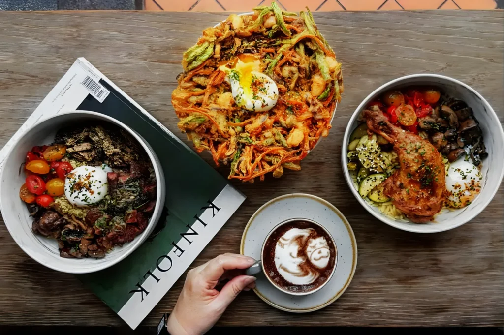 A rustic wooden table with three bowls of vibrant food, each topped with a poached egg. There's a latte with frothy art, and a hand holds the cup.