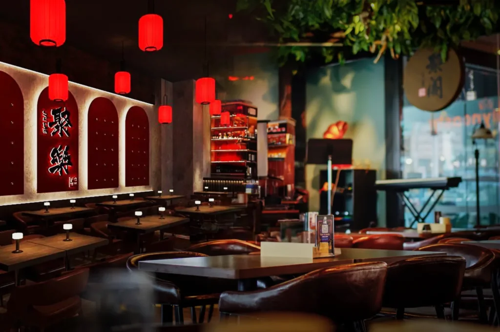 A cozy restaurant with red lanterns, wooden tables, and soft lighting. Walls feature decorative red panels. The ambiance is warm and inviting.