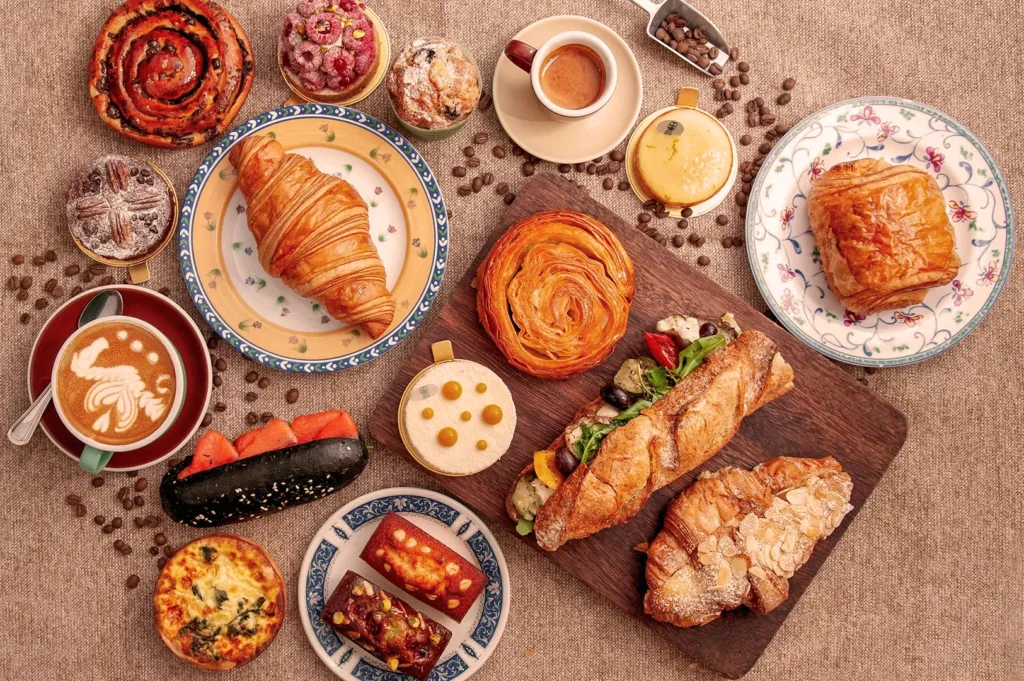 A variety of pastries and breads, including croissants, tarts, and sandwiches, are artfully arranged on a table with cups of coffee, creating a cozy café vibe.