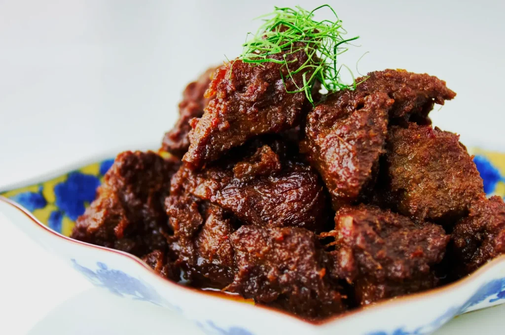 A dish of Rendang, featuring tender, spiced beef chunks garnished with fresh green herbs, served in a blue and white bowl with a floral pattern.