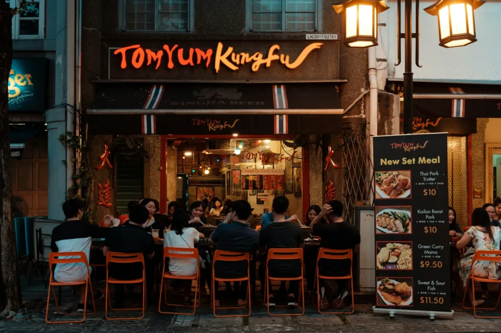 A group of people dine at outdoor tables of a cozy restaurant named "Tom Yum Kungfu." Warm lighting and a menu board create an inviting atmosphere.