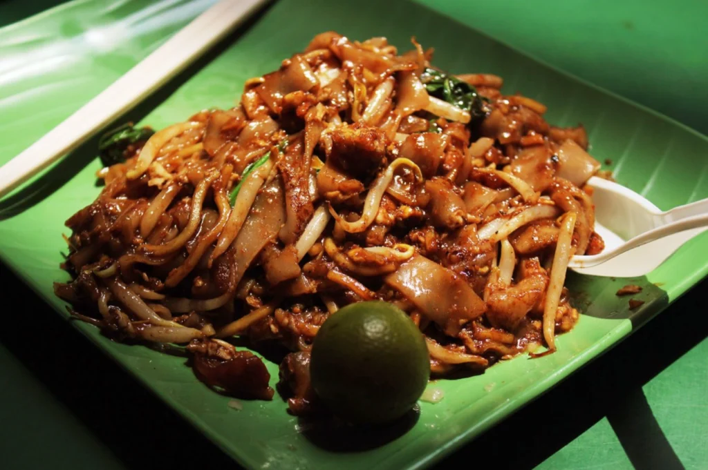 A vibrant plate of char kway teow on a green banana leaf, topped with bean sprouts and accompanied by a lime, is illuminated warmly, evoking a savory delight.