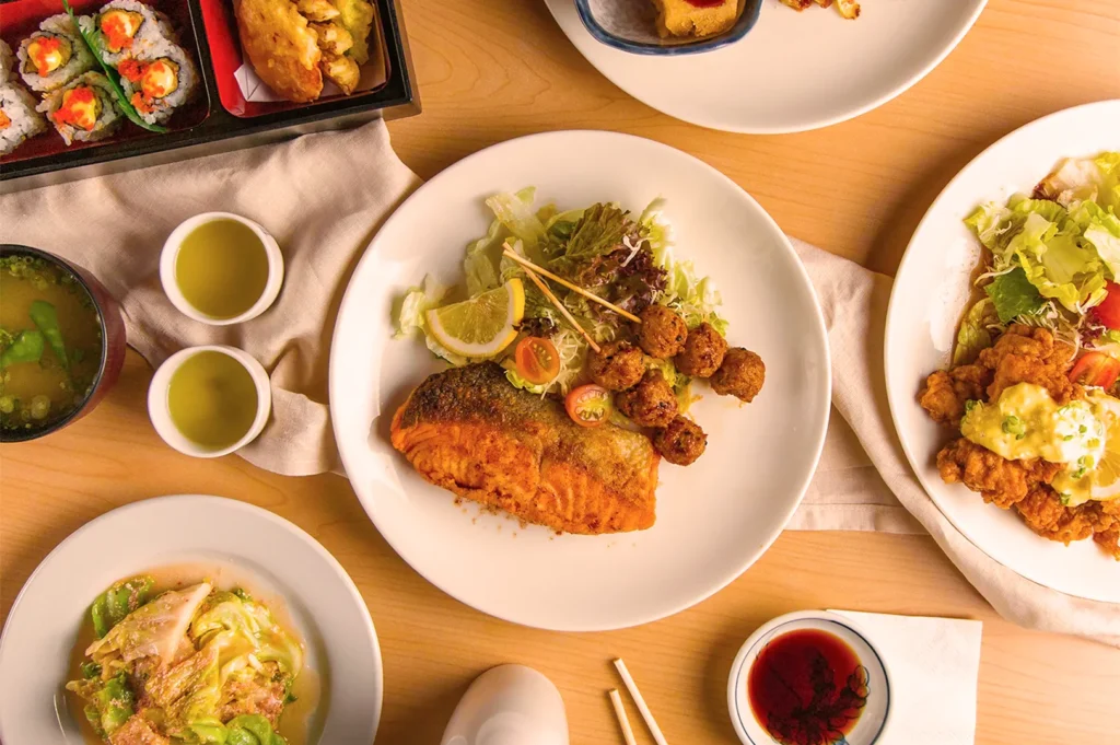 A wooden table features a variety of Japanese dishes, including sushi, salmon, and fried chicken. Green tea, soup, and sauce add to the inviting atmosphere.