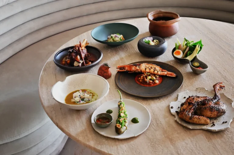 A round table displays an elegant spread of dishes, including grilled meat, seafood, a stuffed chili, boiled egg, and vegetables, evoking a gourmet feast.