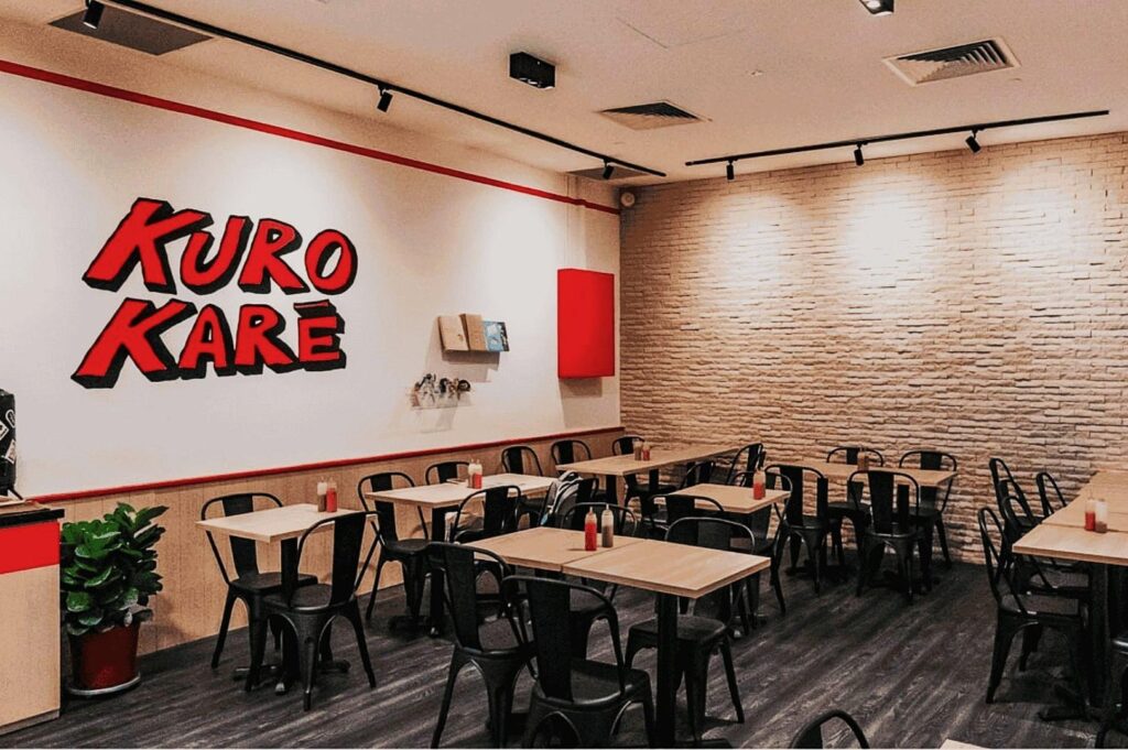 Modern restaurant interior with a minimalist design. White walls feature bold red "Kuro Kare" lettering. Black chairs, wooden tables, and a potted plant create a cozy yet vibrant atmosphere.