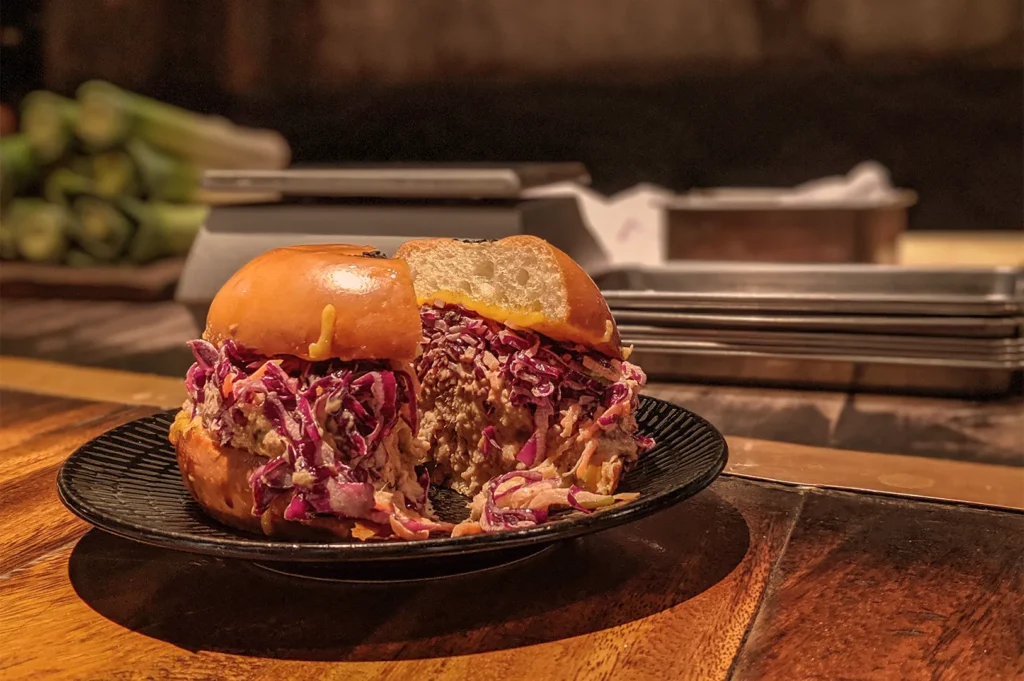 A sandwich with brioche bun filled with colorful coleslaw sits on a black plate. The setting is a warm, rustic kitchen with blurred background items.