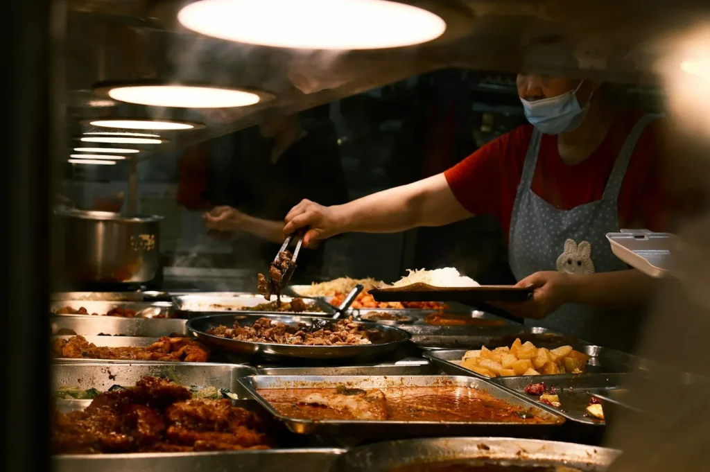 A person wearing a mask and apron serves food from a buffet of various dishes in metal trays, illuminated warmly, suggesting a bustling canteen.