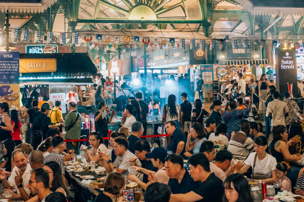 Crowded food market bustling with people enjoying meals, vibrant lights, and smoky stalls; festive atmosphere with diverse, lively crowd.