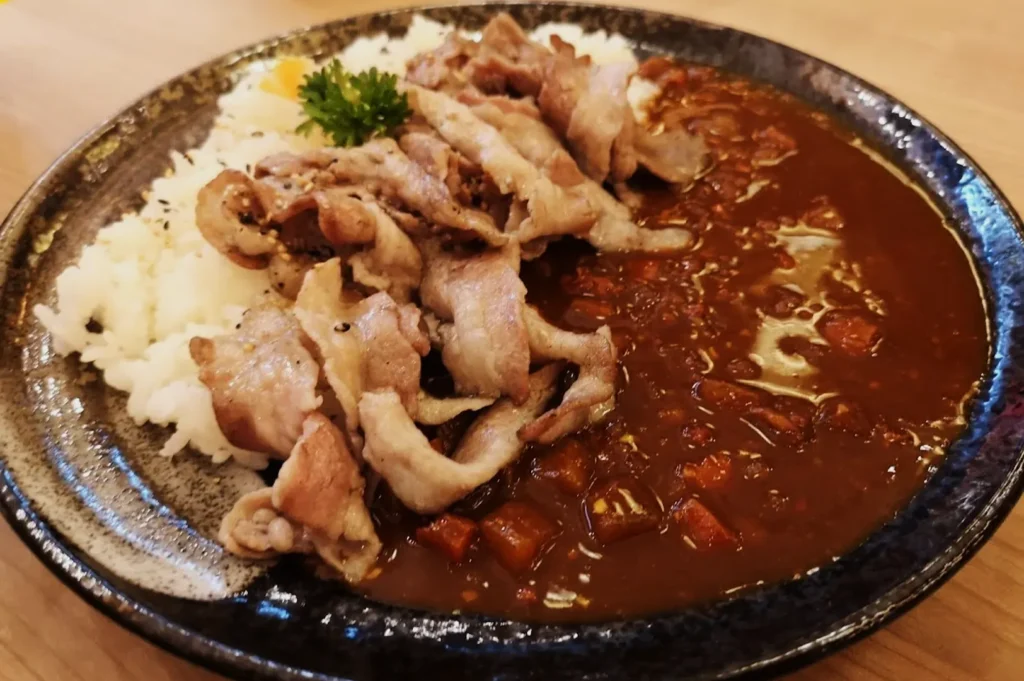 A black plate with sliced grilled meat, white rice topped with parsley, and rich brown curry, creating a hearty and appetizing meal.