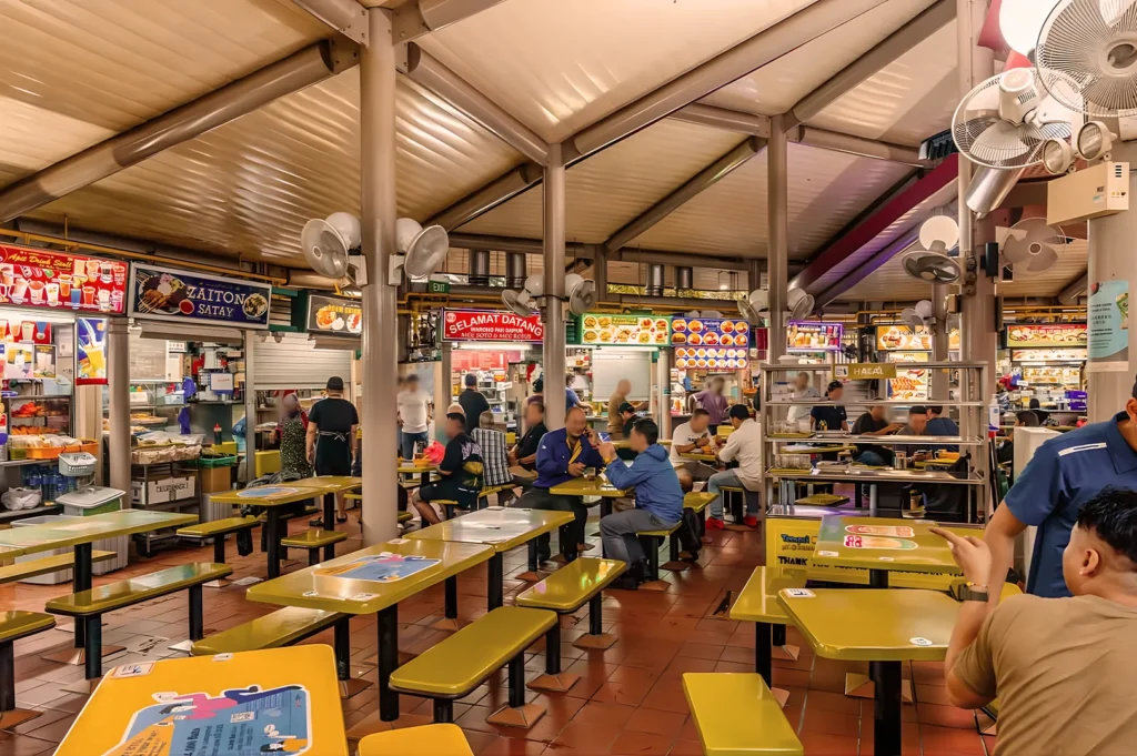 Bustling food court with yellow tables, diverse diners, and colorful food stalls in the background. The atmosphere is lively and vibrant.