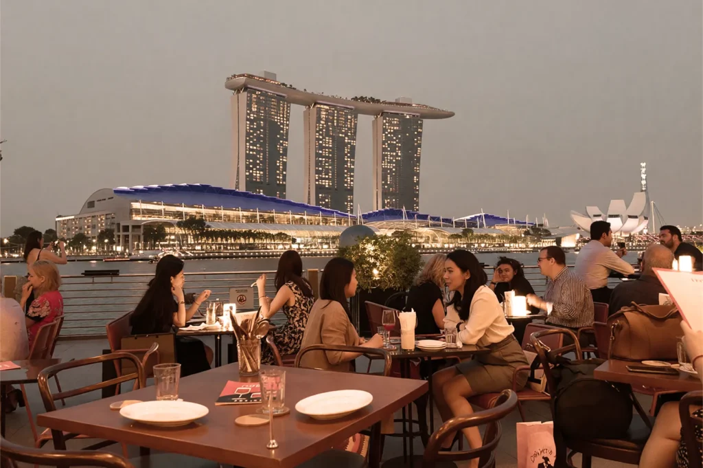 People dining at an outdoor restaurant overlooking Marina Bay Sands in Singapore at dusk. The scene is lively and the sky is softly lit.