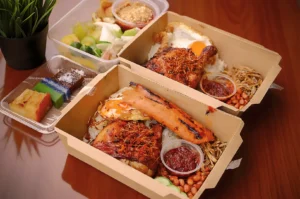 Boxes of nasi lemak on a wooden table feature grilled chicken, sausage, egg, sambal, peanuts, and anchovies. Side dishes include mixed fruit and colorful kuih desserts.