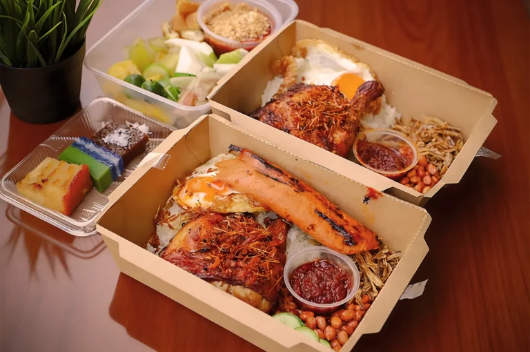 Boxes of nasi lemak on a wooden table feature grilled chicken, sausage, egg, sambal, peanuts, and anchovies. Side dishes include mixed fruit and colorful kuih desserts.
