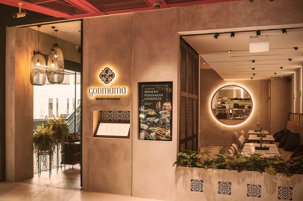 Entrance of a modern Peranakan restaurant with warm lighting, stylish hanging lamps, and a glowing circular mirror. The sign reads "godmama."