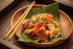 Stir-fried noodles with shrimp and vegetables on a banana leaf, served in a wooden bowl with yellow chopsticks. The setting is warm and inviting.