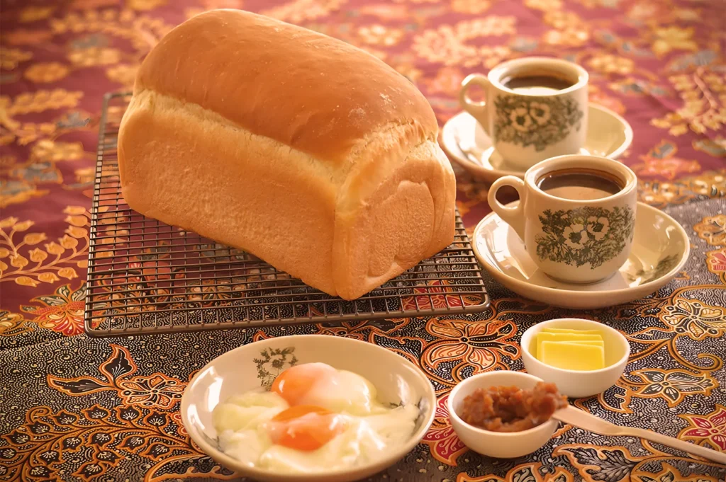 A loaf of bread on a cooling rack, served with soft-boiled eggs, coffee in floral cups, butter, and jam on a vibrant, patterned tablecloth. Cozy and inviting.