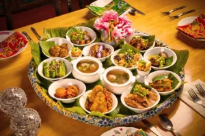 A traditional Thai meal with assorted dishes in small bowls on a banana leaf-lined platter. Brightly colored plates and a pink flower adorn the wooden table.