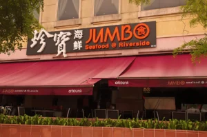 Outdoor view of a seafood restaurant with a large sign reading "JUMBO Seafood @ Riverside" in red and orange. Red awnings cover the seating area.