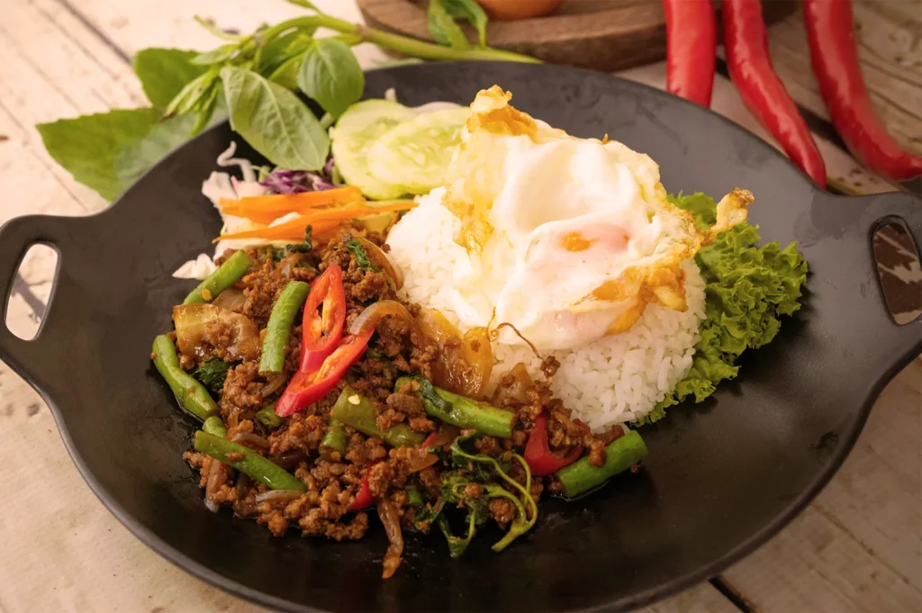 Authentic Thai Basil Minced Pork (Pad Kra Pao) served with white rice, a crispy fried egg, and fresh cucumber garnish on a black plate.