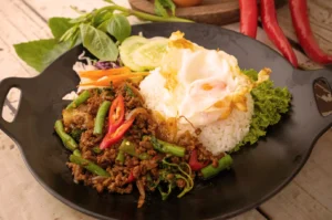 Authentic Thai Basil Minced Pork (Pad Kra Pao) served with white rice, a crispy fried egg, and fresh cucumber garnish on a black plate.