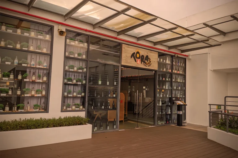 Contemporary restaurant exterior with glass-front windows displaying bottles and plants. Warm tones and sleek design create a welcoming atmosphere.