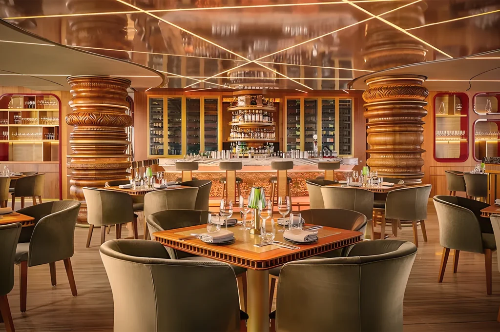 Luxurious restaurant interior with warm lighting, featuring wooden columns, elegantly set tables, plush green chairs, and a stocked bar in the background.