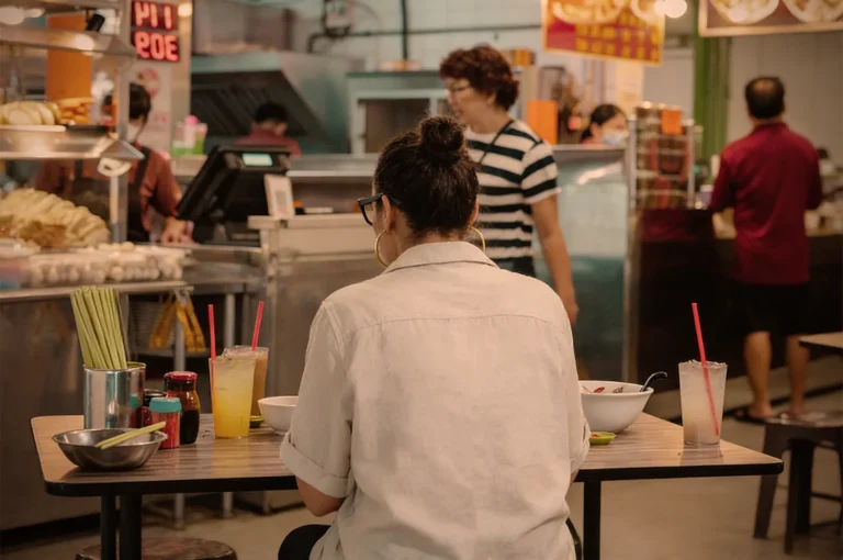 A woman sits at a table in a bustling food court with drinks and noodles. Vendors and other patrons are visible in the background, creating a lively atmosphere.