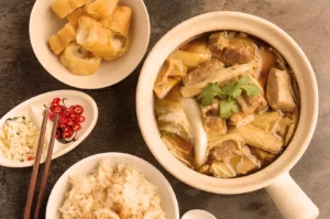 A hearty bowl of bak kut teh with pork ribs and herbs, garnished with cilantro. Accompanied by white rice, fried dough, and chopped garlic with chili slices.