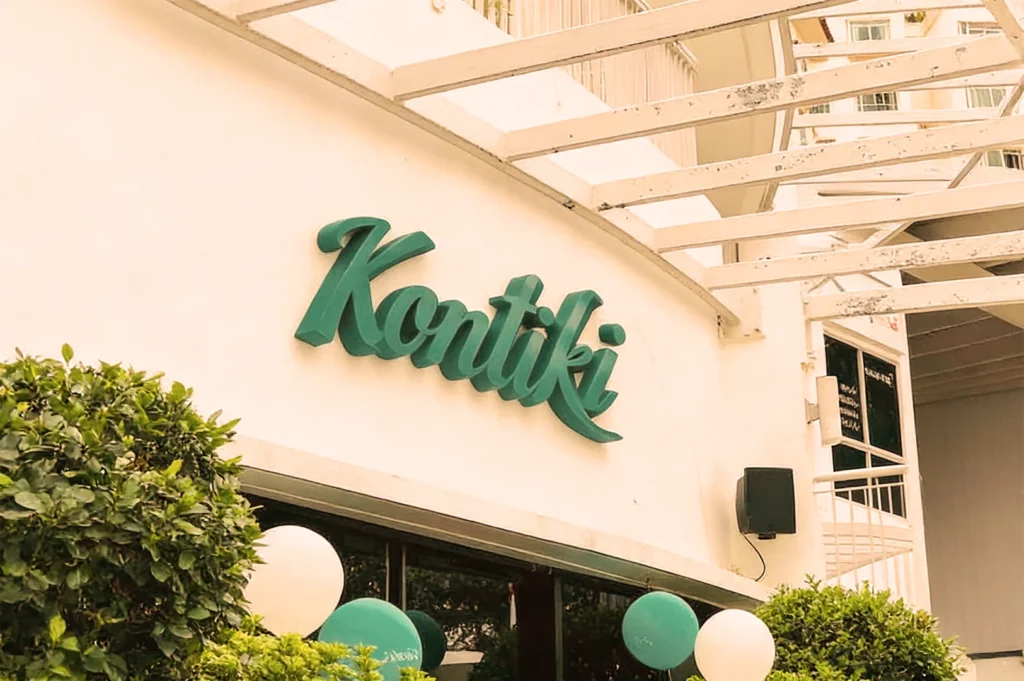 A white building with an overhang features the teal sign 'Kontiki.' Surrounded by green and white balloons, and lush bushes create a festive atmosphere.