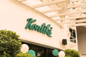 A white building with an overhang features the teal sign 'Kontiki.' Surrounded by green and white balloons, and lush bushes create a festive atmosphere.