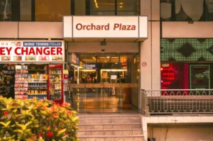 Entrance of Orchard Plaza with illuminated sign; adjacent is a currency exchange booth. The scene is bustling, with vibrant lights and a warm ambiance.