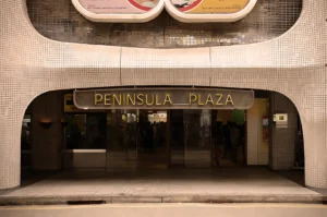 Entrance of Peninsula Plaza with a retro-style sign, tiled facade, and glass doors. Soft lighting creates a vintage, welcoming atmosphere.