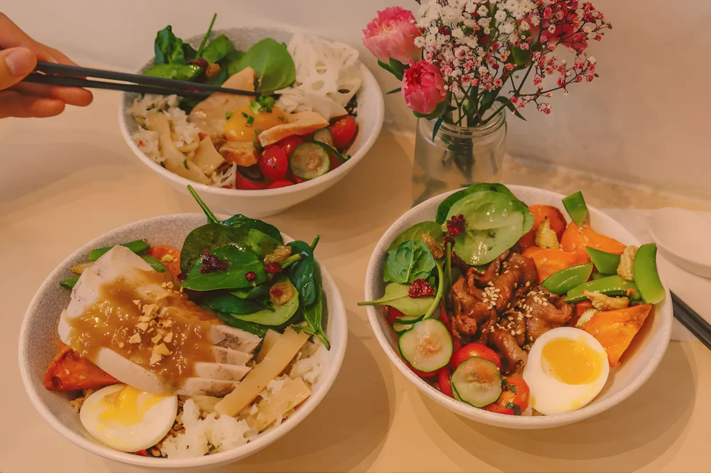 Three colorful rice bowls filled with fresh vegetables, eggs, and protein are placed on a light table. A hand with chopsticks reaches for one bowl. A vase of pink flowers sits nearby, adding a vibrant touch.