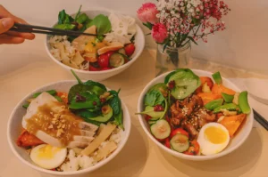 Three colorful rice bowls filled with fresh vegetables, eggs, and protein are placed on a light table. A hand with chopsticks reaches for one bowl. A vase of pink flowers sits nearby, adding a vibrant touch.