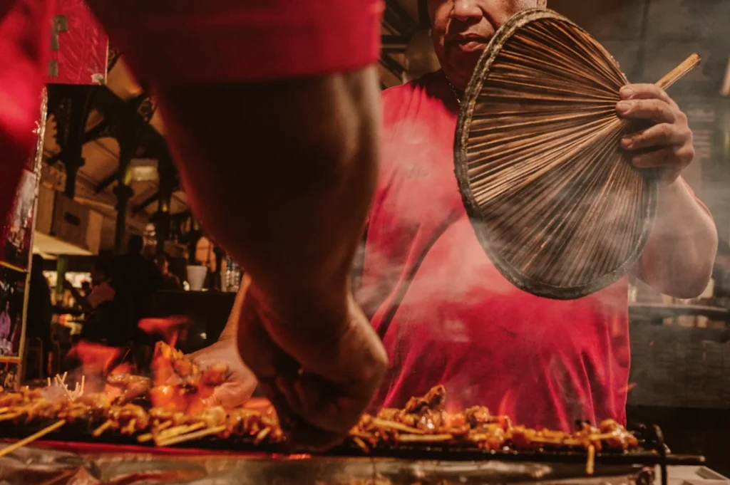 Street food vendor grills skewers, fanning smoke with a round bamboo fan. Warm lighting and bustling market ambiance create a lively, aromatic scene.