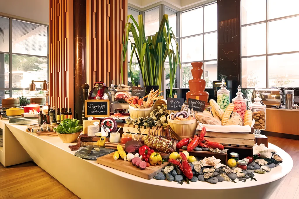 A lavish buffet with a variety of cheeses, breads, and seafood. A chocolate fountain, pastries, and fresh fruits are displayed in a sunny, elegant setting.