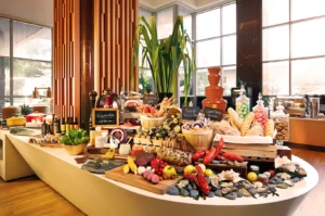 A lavish buffet with a variety of cheeses, breads, and seafood. A chocolate fountain, pastries, and fresh fruits are displayed in a sunny, elegant setting.