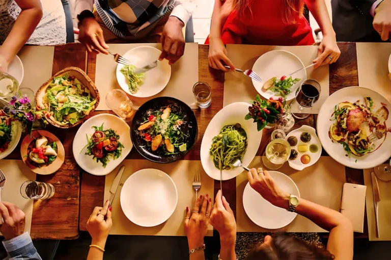 Top-down view of a dining table with seven people sharing a meal. Plates of colorful salads and pasta are surrounded by glasses, creating a lively, communal atmosphere.