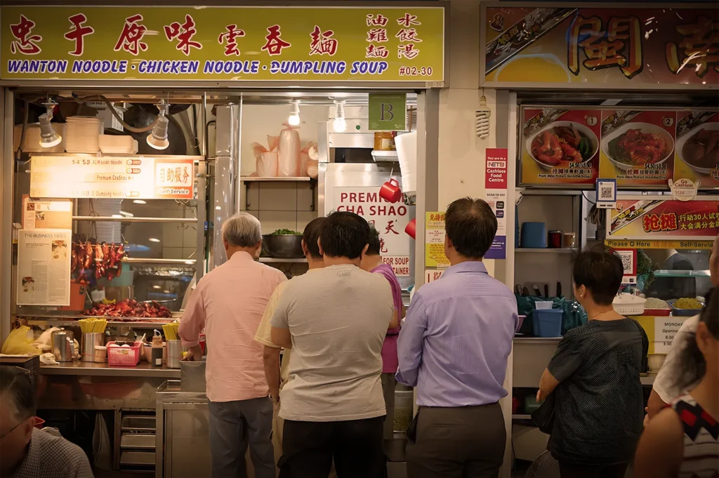 People queue at a bustling noodle stand under a sign reading "Wanton Noodle • Chicken Noodle • Dumpling Soup." The warm lighting creates a lively, inviting atmosphere.