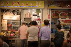 People queue at a bustling noodle stand under a sign reading "Wanton Noodle • Chicken Noodle • Dumpling Soup." The warm lighting creates a lively, inviting atmosphere.
