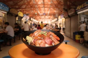 A vibrant seafood rice bowl garnished with salmon and roe is centered on an orange table, set in a bustling hawker center filled with diners.