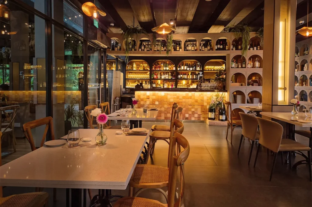 Cozy restaurant interior with warm lighting and wood accents. Tables are set neatly, featuring fresh flowers. A well-stocked bar is in the background.