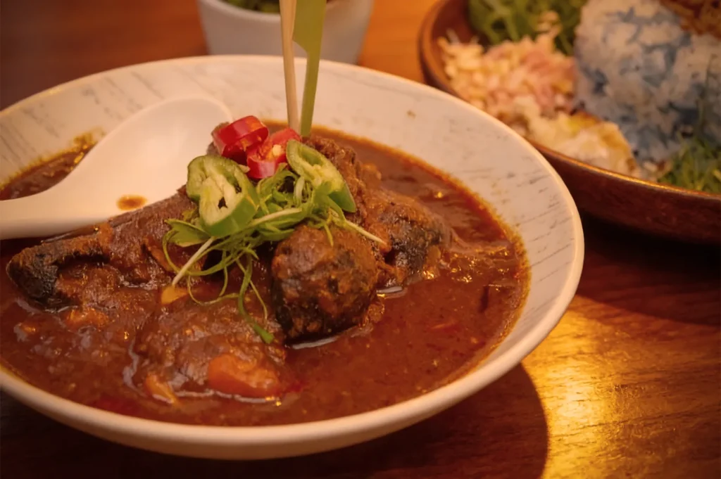 A bowl of rich, dark curry with tender chunks of meat garnished with sliced red and green chilies and herbs, resting on a wooden table.