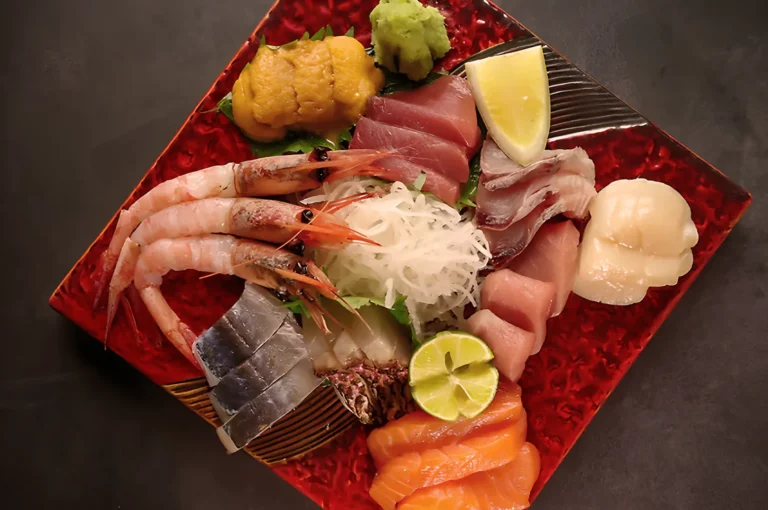 A colorful sashimi platter on a red plate, featuring shrimp, tuna, salmon, scallop, mackerel, and a lime slice, garnished with wasabi and shredded radish.