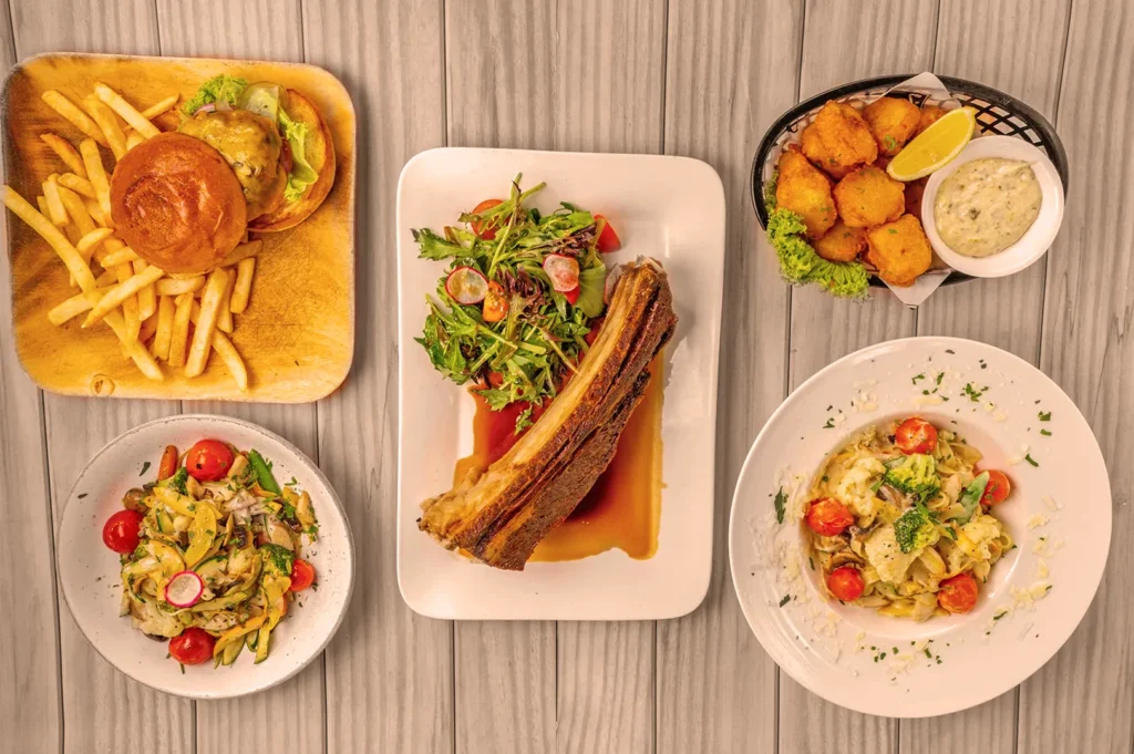 Aerial view of a varied meal on a wooden table, featuring a burger with fries, a pork rib with salad, pasta with tomatoes, and fried appetizers. Luxurious and appetizing.