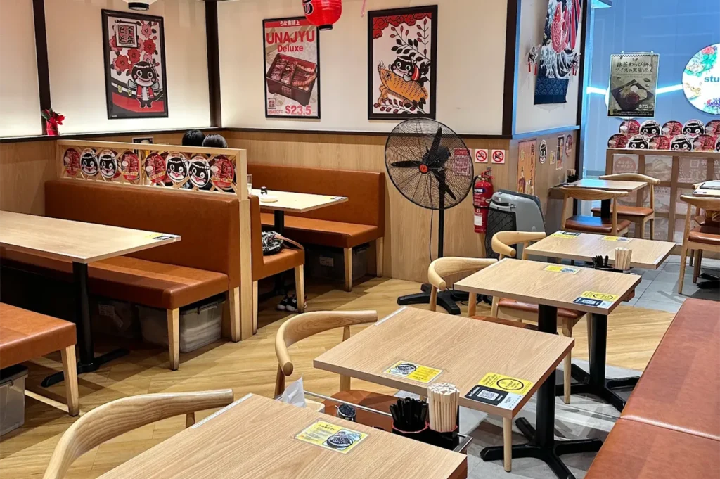 Cozy dining area interior of Unatoto Japanese restaurant with wooden tables, comfortable brown leather booth seating, wall posters, and a large standing floor fan.