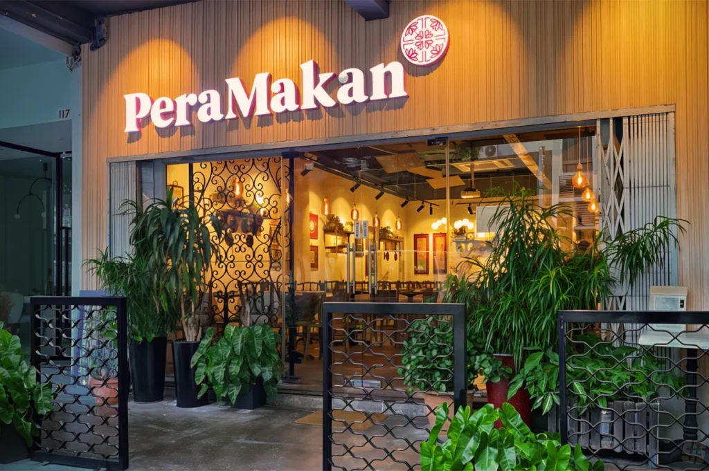Restaurant entrance with "PeraMakan" sign, decorated with potted plants and warm lighting. The glass doors reveal cozy interior seating.