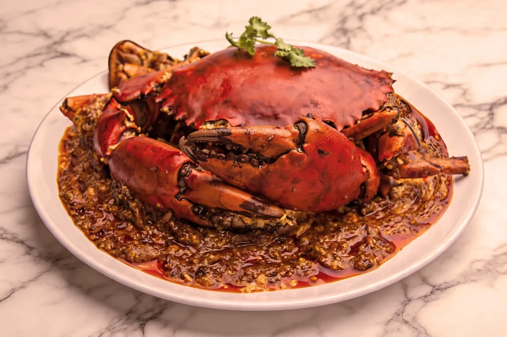 Spicy crab dish presented on a white plate with a rich, red sauce garnished with cilantro, set on a marble surface, evoking a savory aroma.