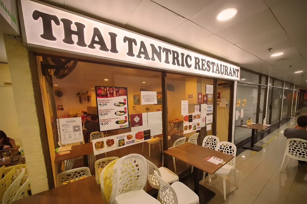 Entrance to Thai Tantric Restaurant featuring a bright lightbox sign, casual indoor seating, and menu posters on the glass windows.