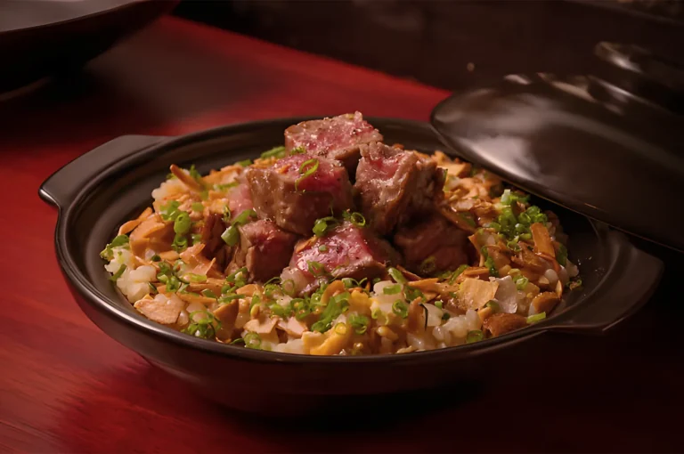 A black ceramic bowl on a wooden table filled with rice, mushrooms, scallions, and cubes of juicy, seared meat. The dish looks savory and comforting.